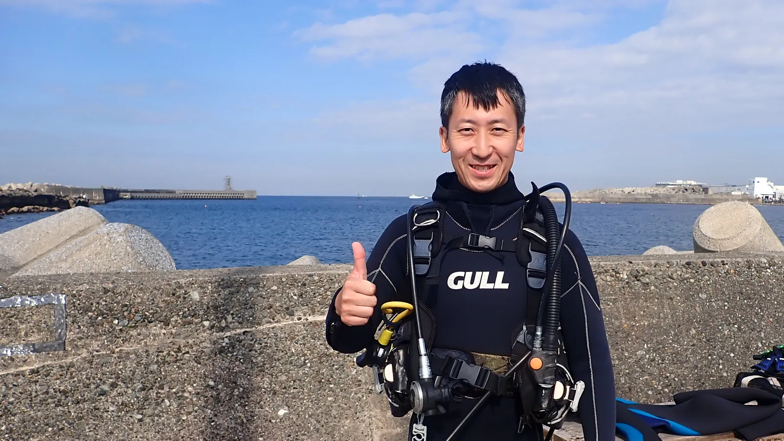 青空の海辺で、ウェットスーツとBCD、レギュレーターを身につけて、笑顔で親指を立てるダイバー